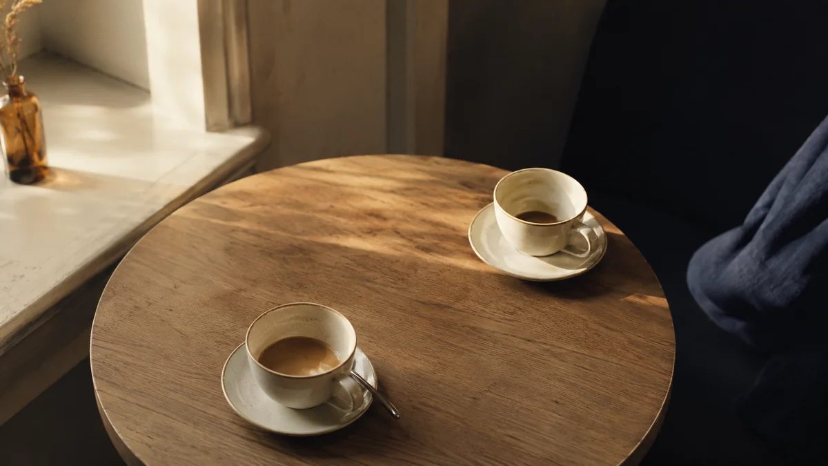 木のテーブルに置かれた二つのコーヒーカップ。ひとつは空に近く端へ寄り、もうひとつはまだ半分残って手前にある。ふたりのあいだに何が残っているかを示す。
