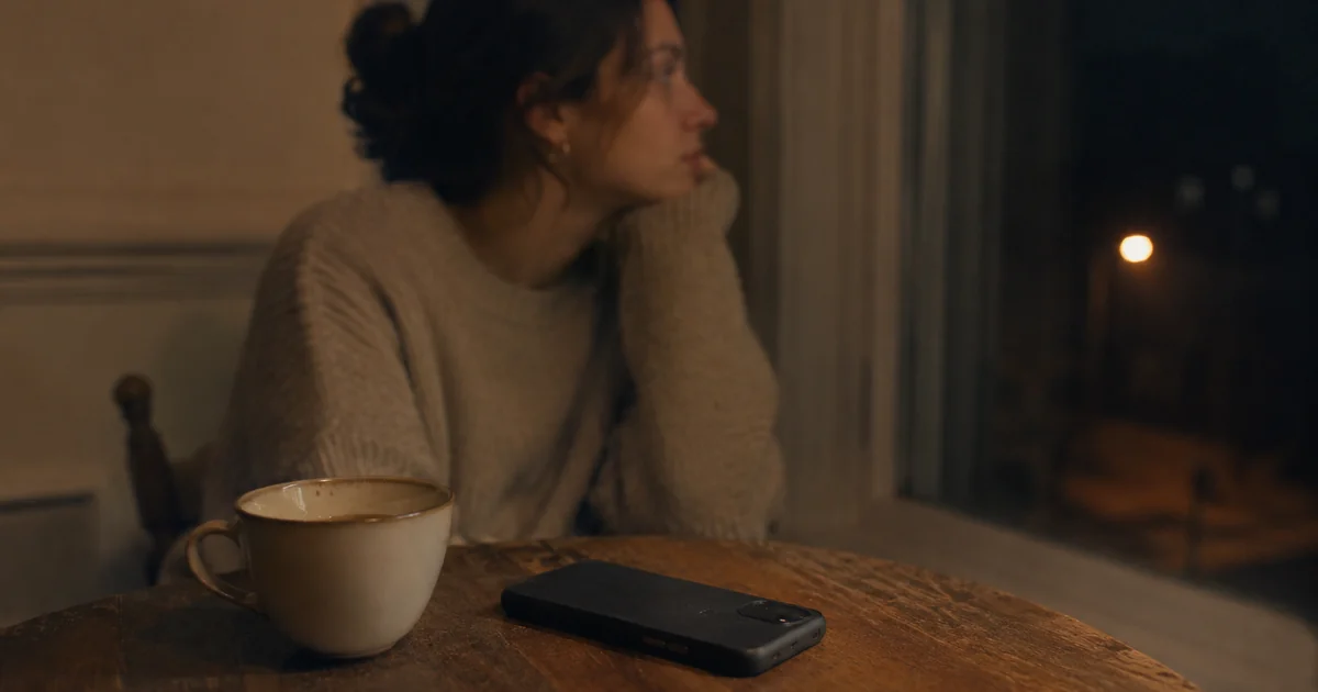 夜のテーブルに伏せて置かれたスマートフォンと飲みかけのカップ。返信が来ないまま時間が過ぎる、ゴースティングの曖昧な沈黙を表す。